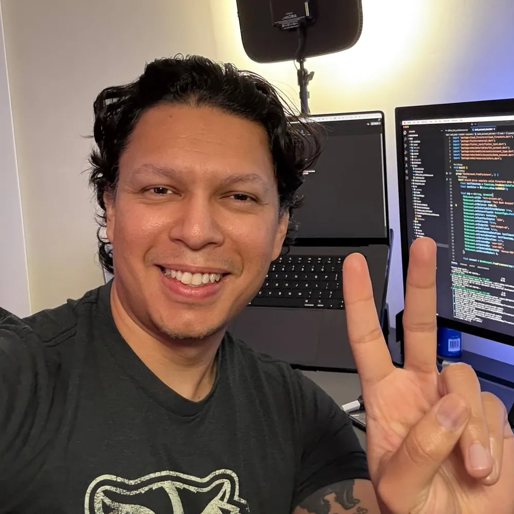 Julio Cruz, founder of NEKO, at a desk with code and the NEKO app on screen, making a peace sign
